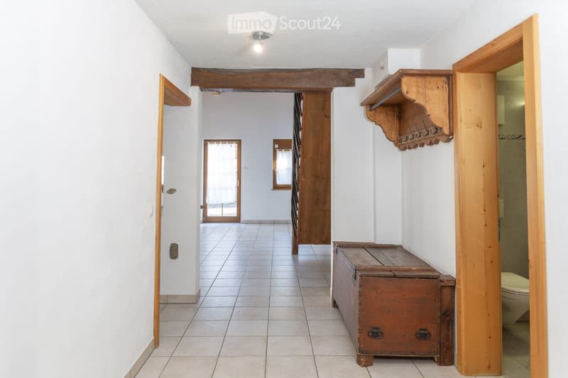 A well-lit hallway with tiled flooring, white walls, and wooden beams. Features include a wooden chest, a coat rack, and an open doorway leading to another room.