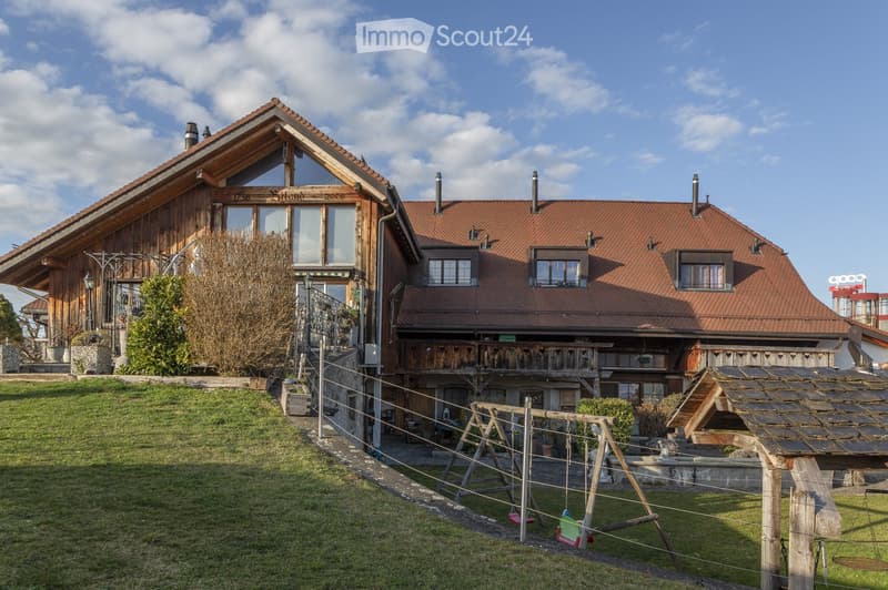 Two-story house with red-tiled roof, wooden balcony, and surrounding garden with swing set.