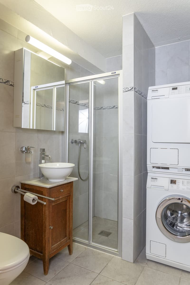 Modern bathroom with a wooden vanity, white sink, mirror, glass-enclosed shower, and a stacked washer/dryer unit.