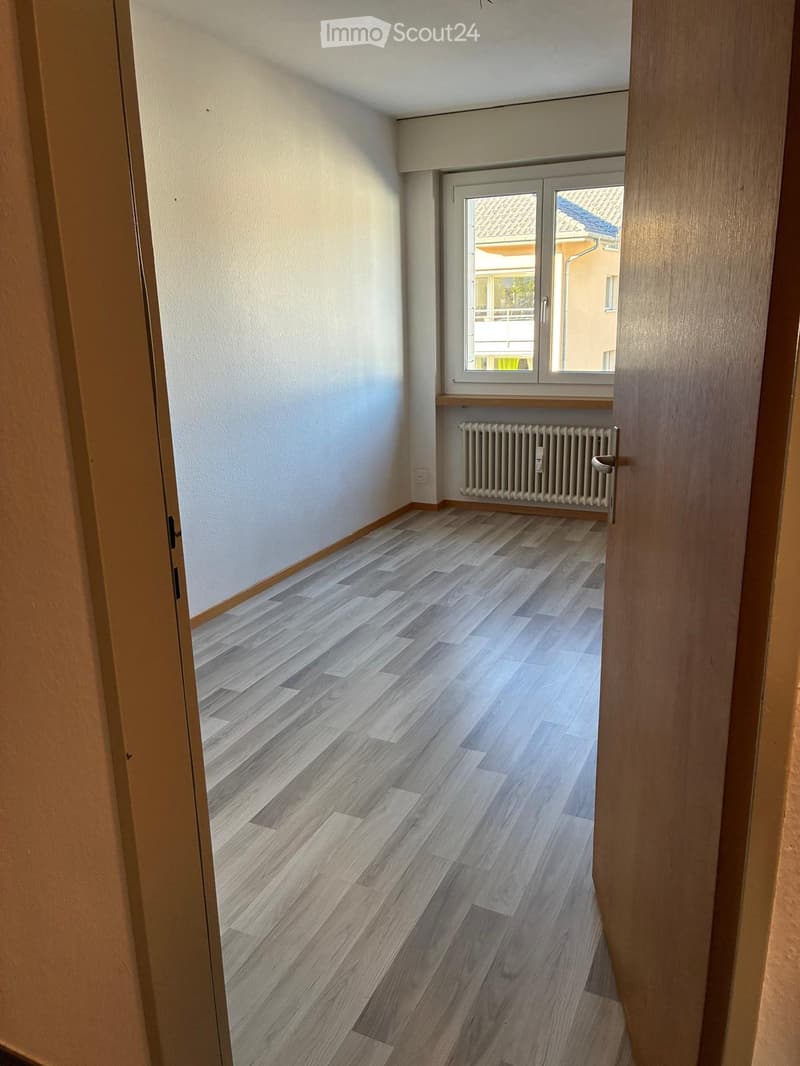 Empty room with light wooden flooring, white walls, a window with a radiator below, and a door frame visible on the left side.