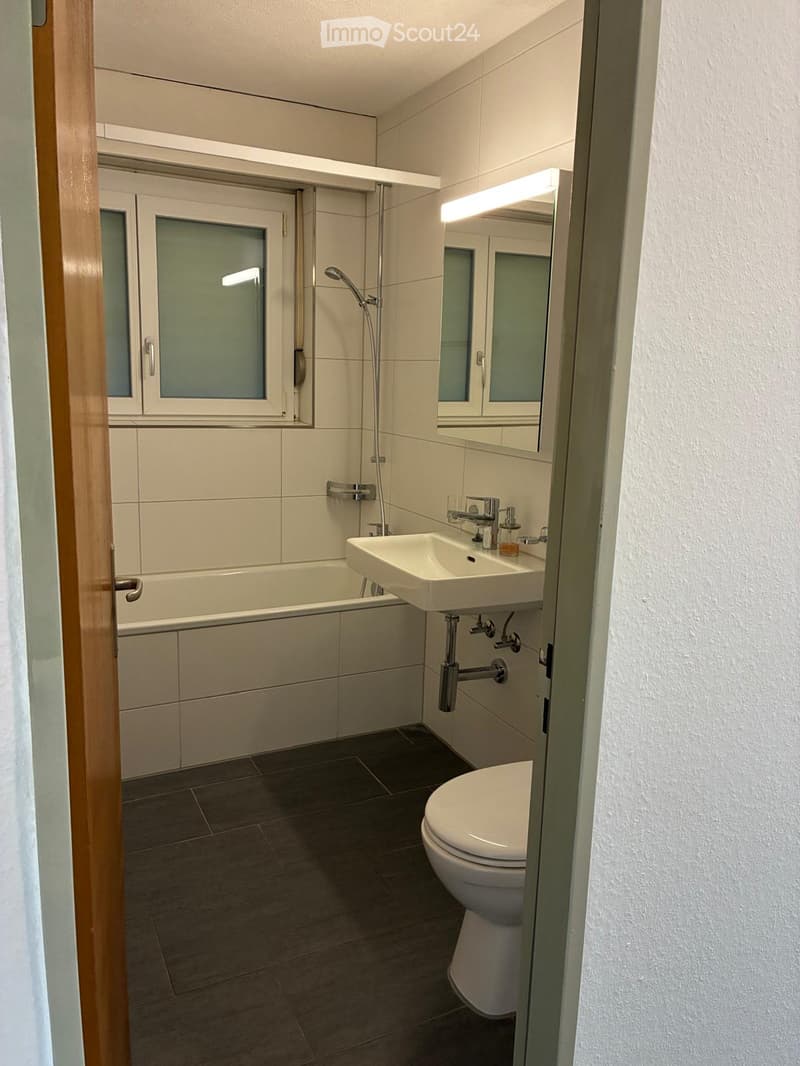 Modern bathroom with a toilet, sink, bathtub, and showerhead. Features a large mirror with built-in lighting and a window with frosted glass.