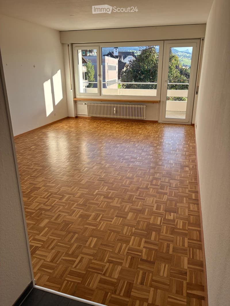 Empty room with wooden herringbone flooring, large windows, radiator, and sliding glass door leading to a balcony.