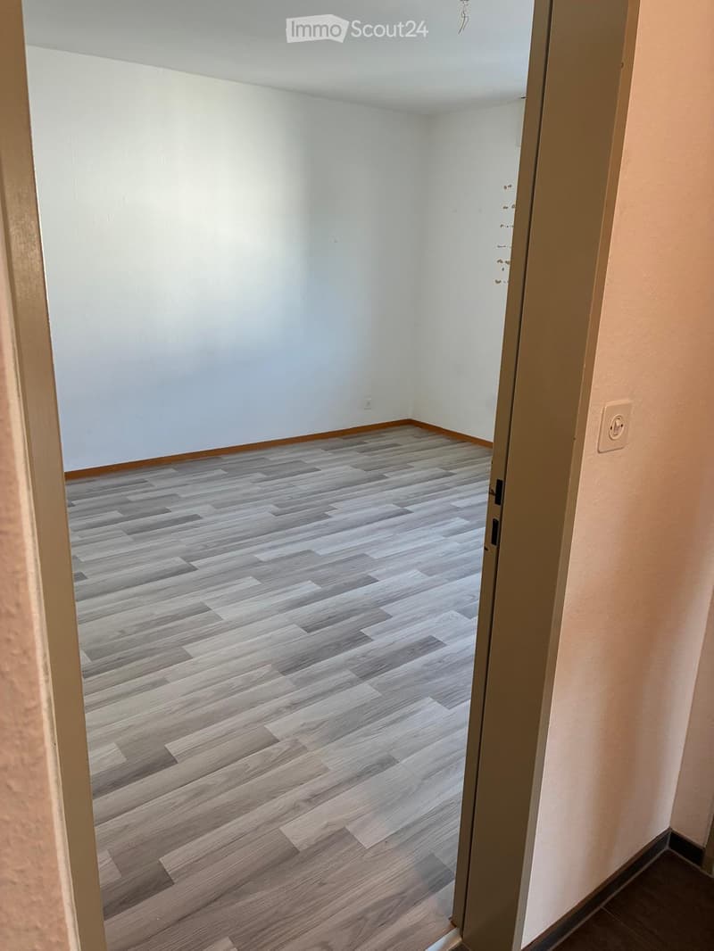 Empty room with light-colored wooden flooring and white walls, viewed from an open doorway.