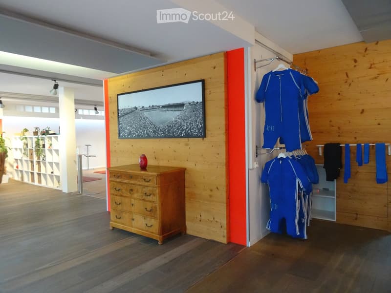 A modern, well-lit room with wooden flooring, a wooden dresser, a large screen mounted on the wall, and several blue sports outfits hanging on a rack. The room features a sleek, contemporary design with a mix of wooden and white elements.