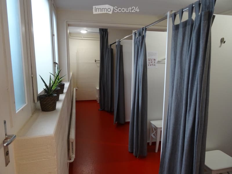 A narrow hallway with red carpeting, white walls, and grey curtains on a rod. There is a small white stool and a white table near the end of the hallway, with a window on the left side showing potted plants on the windowsill.