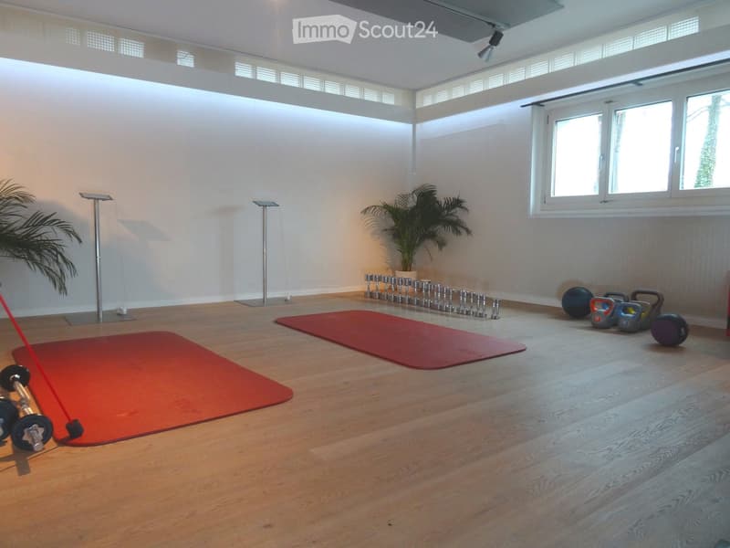 A spacious, well-lit fitness room with wooden flooring, yoga mats, dumbbells, kettlebells, and potted plants.