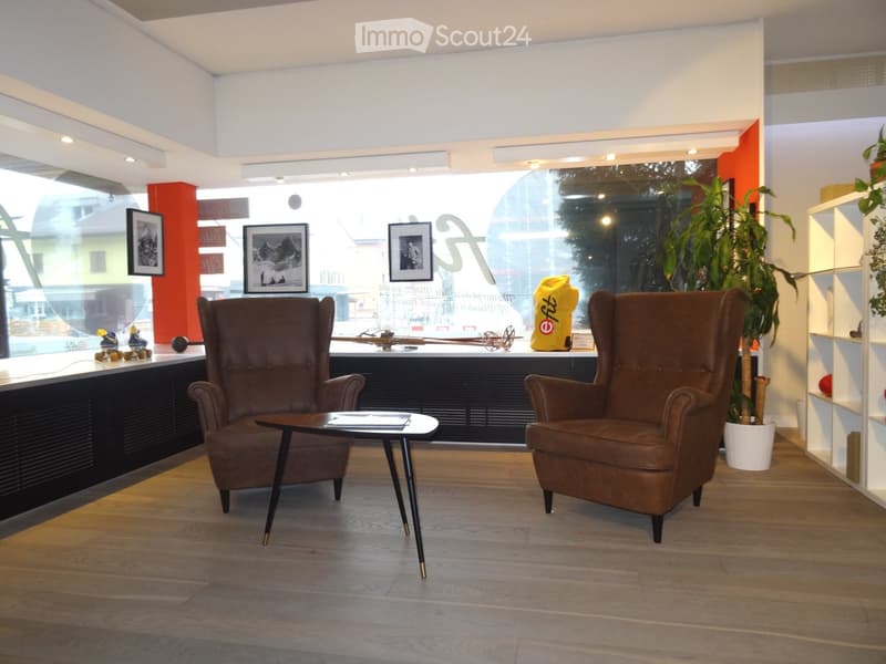 Modern interior with two brown leather chairs, a small black table, framed pictures on the wall, and plants in pots. The room has a polished wooden floor and large windows with a view of the street.