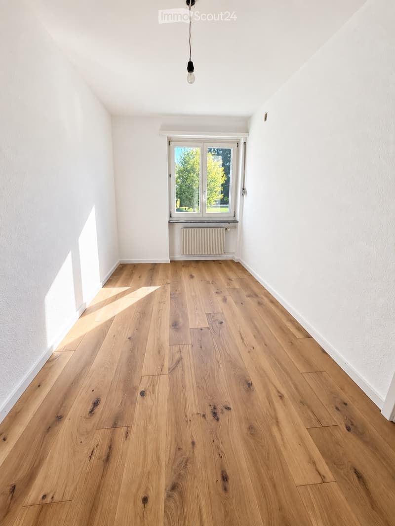 Empty room with light wooden flooring, white walls, and a large window with a radiator below it. Natural light streams in, illuminating the space.