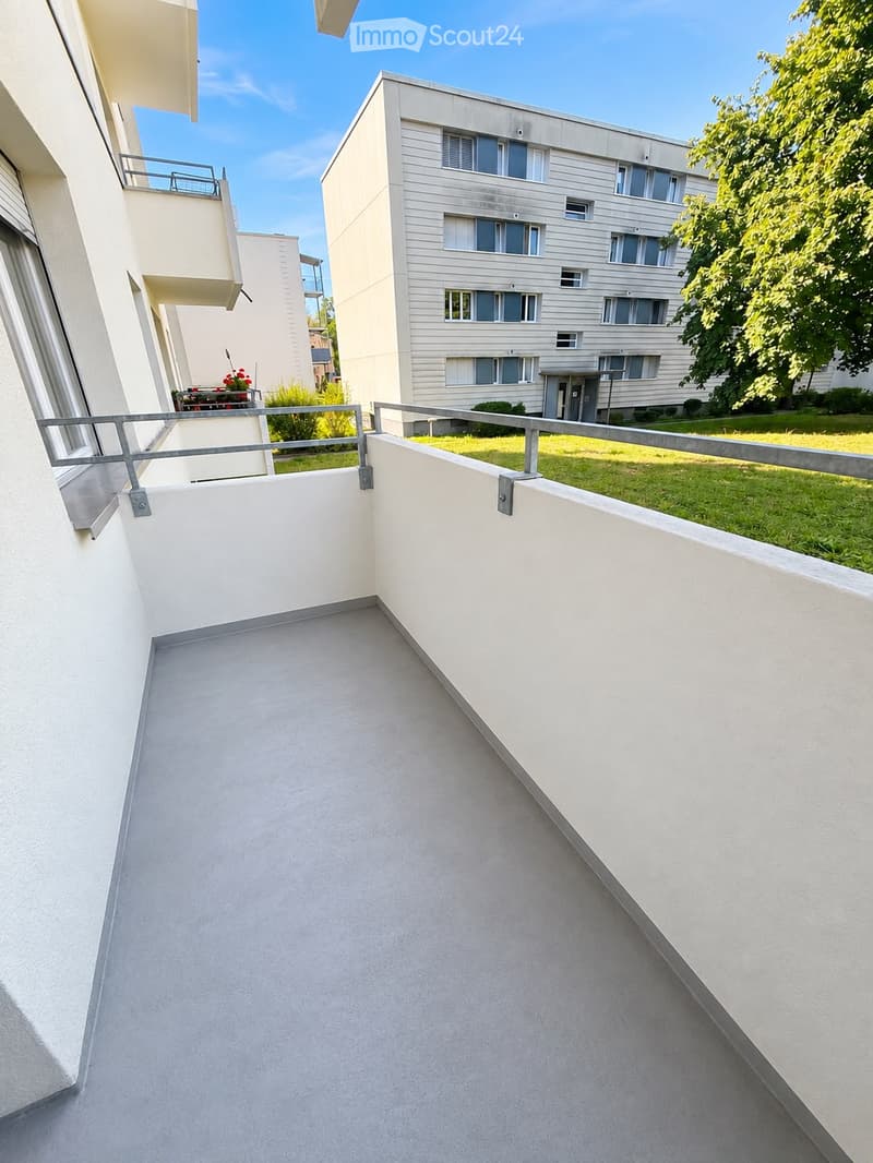 Balcony with white walls and metal railing, overlooking a grassy area and a modern apartment building with multiple windows.