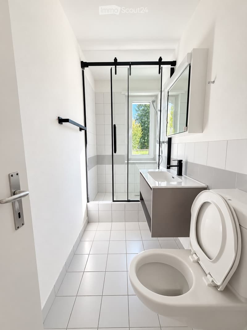 Modern bathroom with white tiles, a toilet, sink, mirror, and a glass-enclosed shower area with a window.
