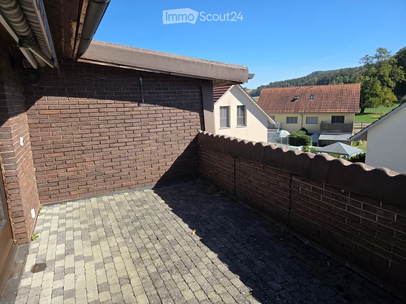 Brick-walled balcony with tiled floor, overlooking neighboring houses and greenery.