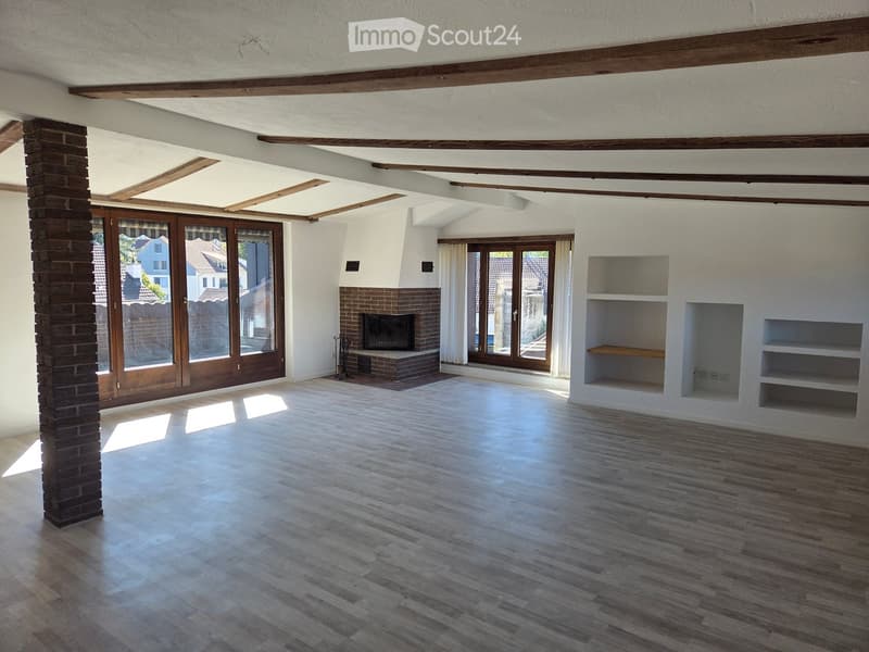 Empty room with wooden flooring, white walls, exposed wooden beams, large windows, and a brick fireplace.