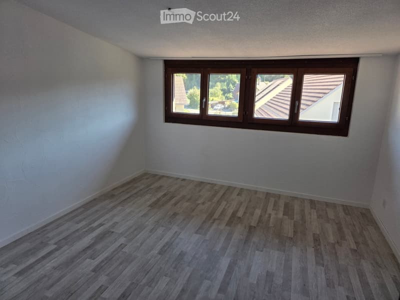 Empty room with light-colored walls and wooden flooring, featuring a large window with brown frames offering a view of neighboring rooftops and greenery.