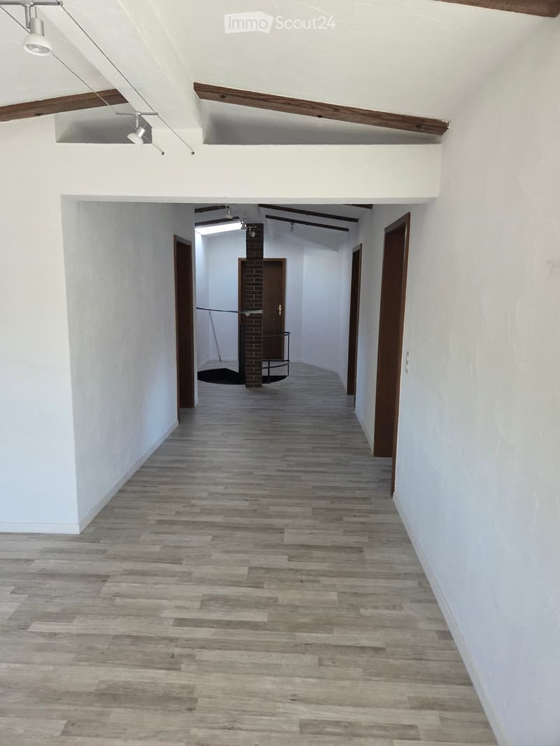 Empty hallway with white walls, wooden flooring, and exposed wooden beams on the ceiling. Features several doors and a small table in the background.