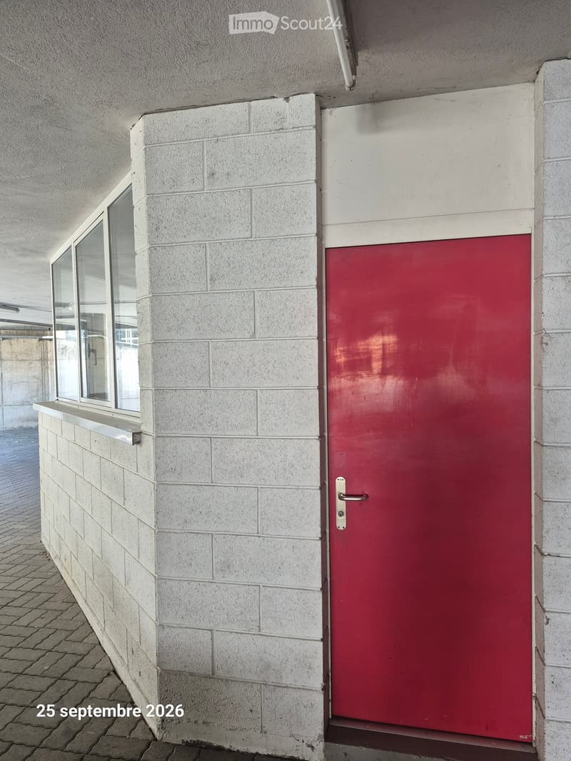 Exterior view of a building entrance with a red door and a window to the left, featuring light-colored brick walls and a tiled pavement.