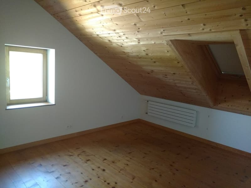 Empty attic room with wooden slanted ceiling, wooden floor, white walls, and a single window letting in natural light.