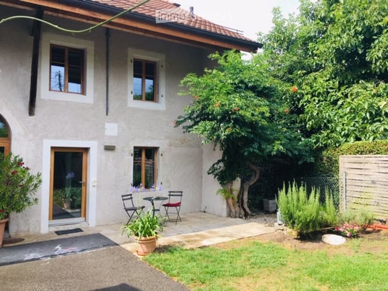 A small, well-maintained garden area adjacent to a house. Features include a patio with a small table and chairs, potted plants, a tree, and neatly trimmed grass.