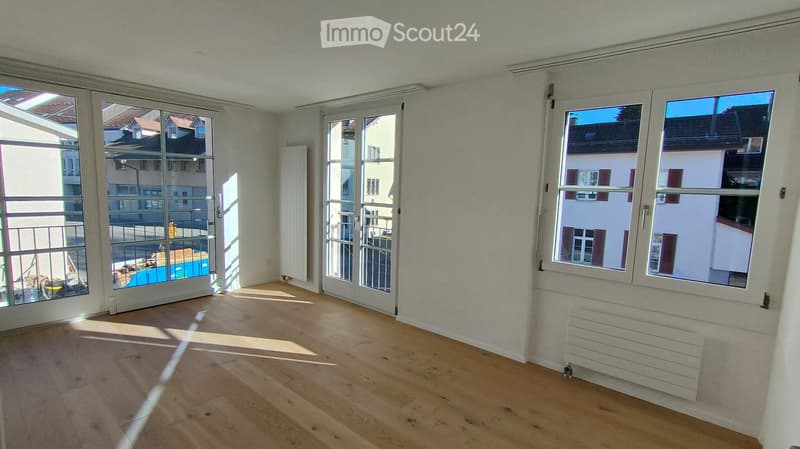 Empty room with wooden flooring, large windows, and sliding glass doors leading to a balcony.