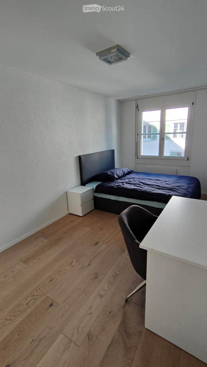 A small bedroom with wooden flooring, a bed with dark bedding, a nightstand, a desk, and a chair.