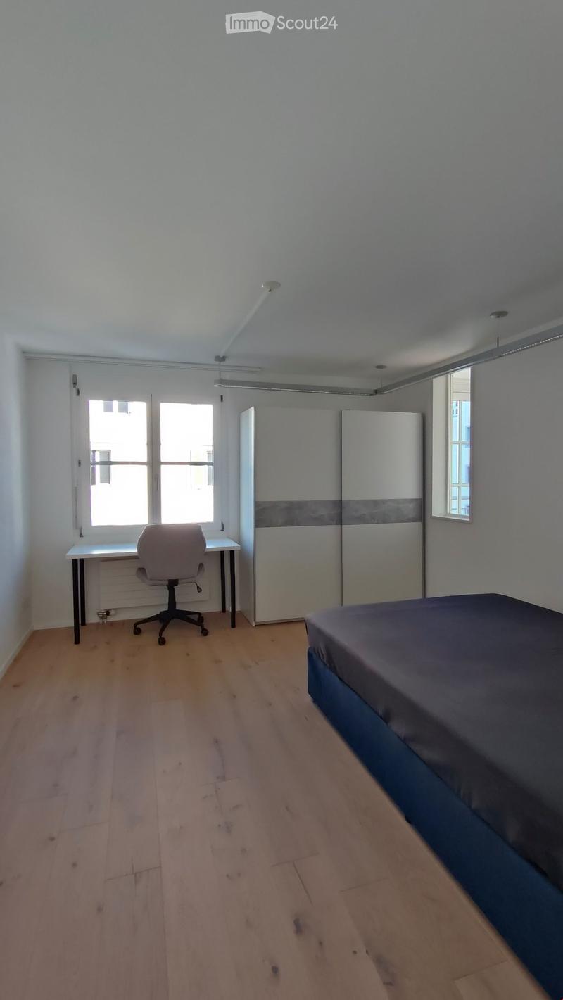 A furnished bedroom with a bed, a desk with a chair, and a wardrobe. The room has wooden flooring and large windows.