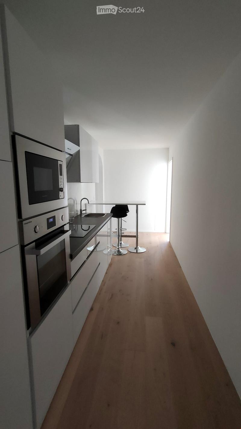 Modern kitchen with light wooden flooring, white cabinets, microwave, oven, sink, and two stools at a small table.