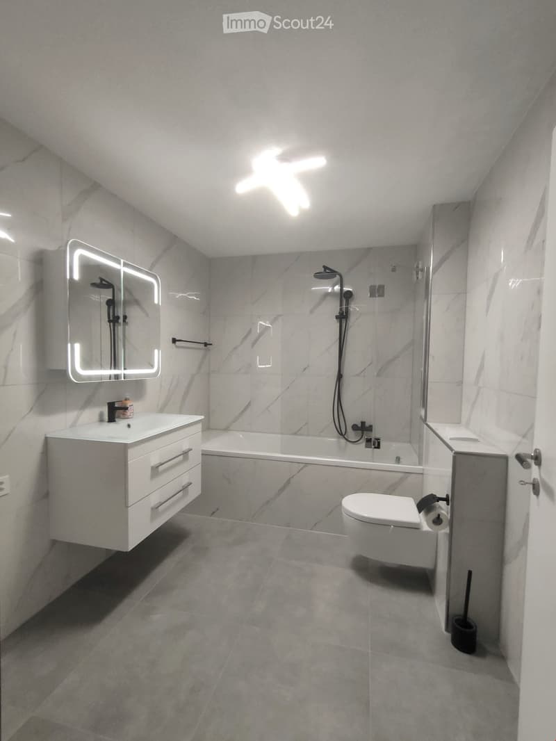 Modern bathroom with marble tiles, a white sink with a mirror, a bathtub, and a toilet. Features include a sleek showerhead and a built-in storage cabinet.