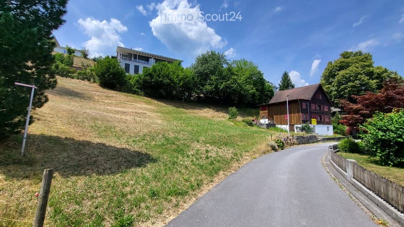 A scenic view of a residential area with a raised ground floor house on a hillside, surrounded by greenery and trees.