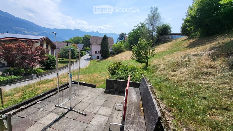 Roof terrace with tiled surface, metal fence, and surrounding greenery, overlooking a residential area with houses and mountains in the background.
