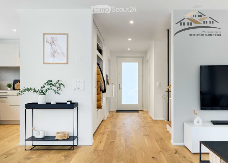 Modern corridor leading to the living room with hardwood floors and white walls, illuminated by recessed ceiling lights, a TV on a side table, a console table with a plant, a picture frame on the wall, an entrance door with a glass panel, and an open ward