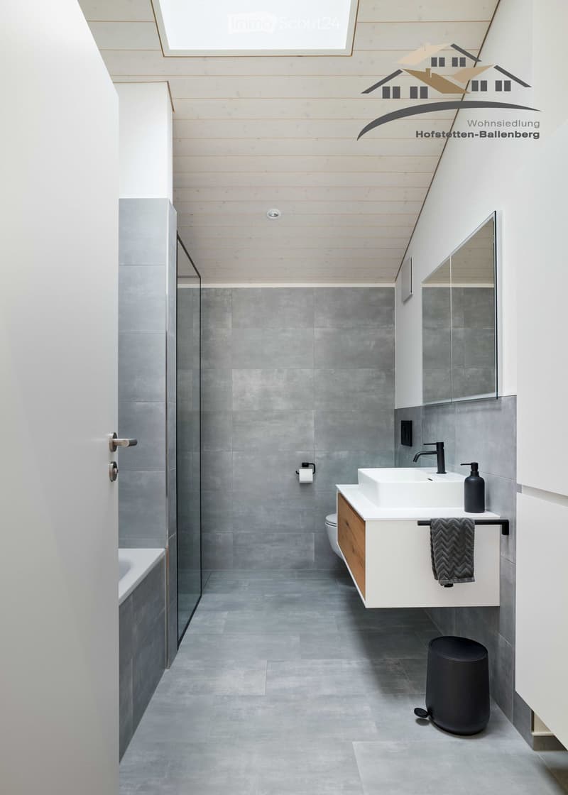 A modern bathroom featuring gray tiled walls, a walk-in shower with a glass door, a white bathtub, a wooden cabinet with a sink, and a mirror above the sink.