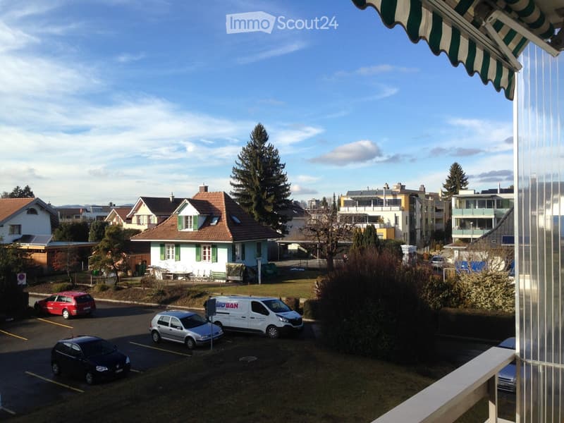 View from a balcony showing a parking area with several cars, a small park with benches and tables, and residential houses in the background under a clear sky.