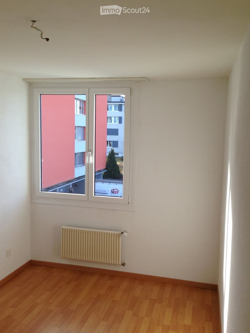 Empty room with wooden flooring, white walls, a radiator, and a large window offering a view of neighboring buildings.