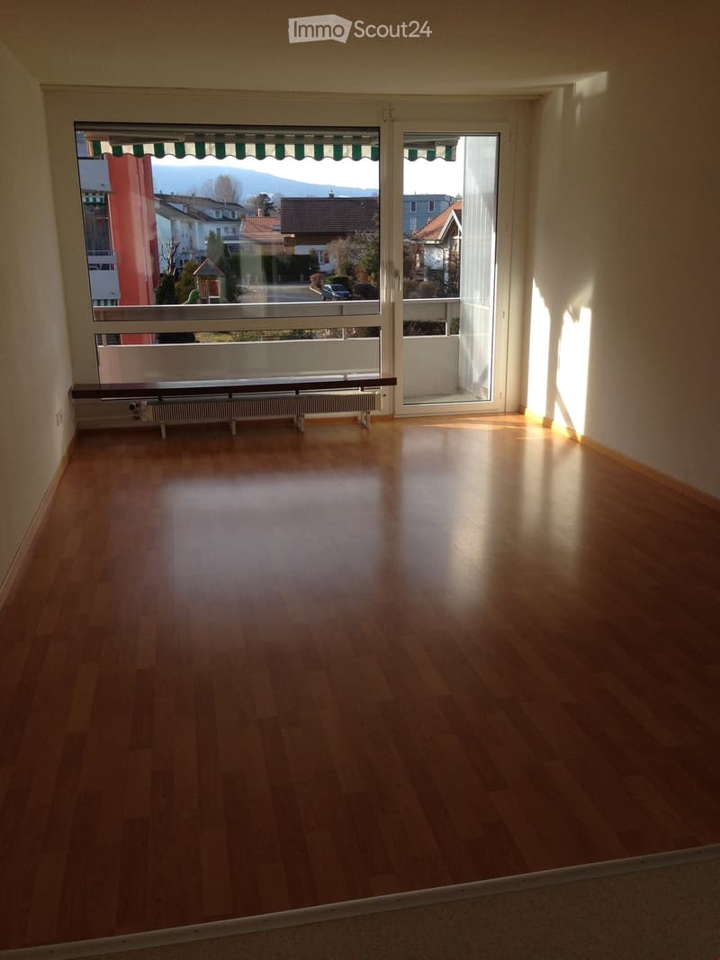 Empty room with wooden flooring, large windows, radiator, and a view of the street and surrounding buildings.