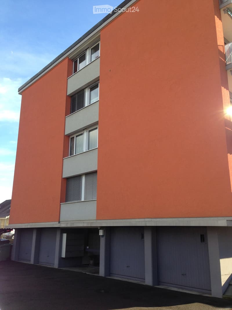 Modern multi-story building with orange facade, white window frames, and balconies. Features a covered parking area with grey garage doors.