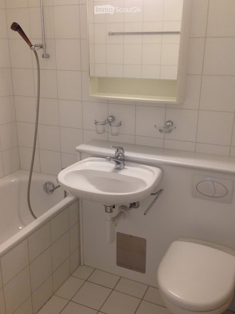 A small bathroom with a white tiled wall and floor, featuring a sink with a faucet, a bathtub with a shower hose, a toilet, a mirror, and a soap dish.