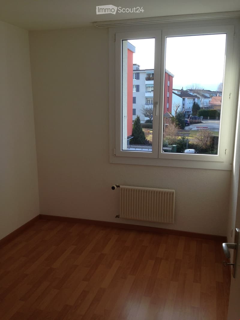 Empty room with wooden flooring, white walls, a radiator, and a large window offering a view of the street and surrounding buildings.