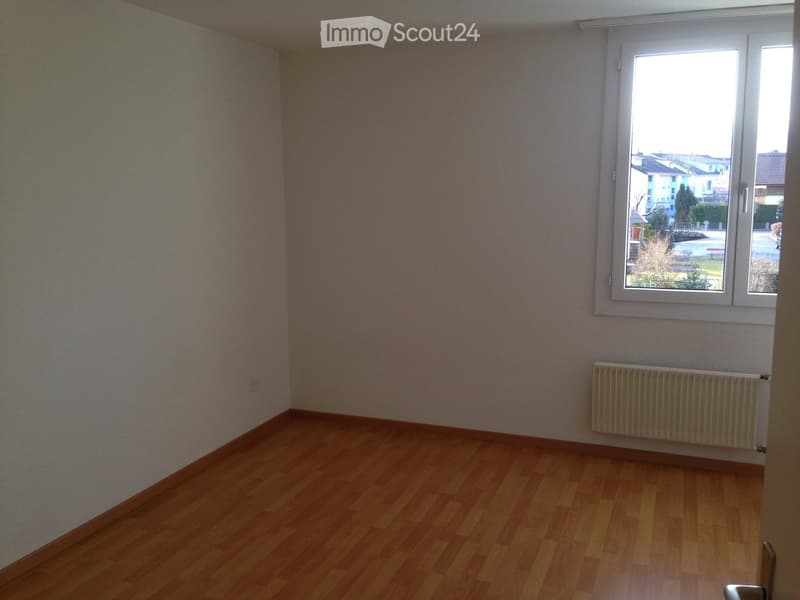Empty room with wooden flooring, light-colored walls, and a window offering a view of the outside. A radiator is visible under the window.