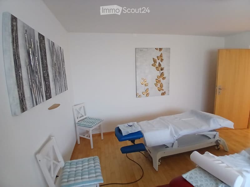 A small, well-lit room with a treatment table, two chairs, and artwork on the walls.