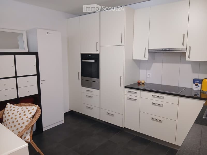 Modern kitchen with white cabinets, stainless steel appliances, black countertops, and a wicker chair with a patterned cushion.