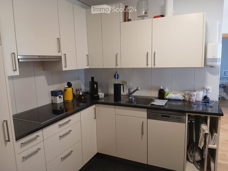 Modern kitchen with white cabinets, black countertops, and stainless steel appliances including an oven, coffee maker, and toaster. A sink with a faucet is visible, along with a dishwasher and various small items on the counter.