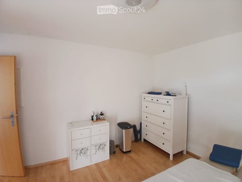 A furnished room with a bed, dresser, nightstand, and a small trash can. The room has light wooden flooring and white walls.