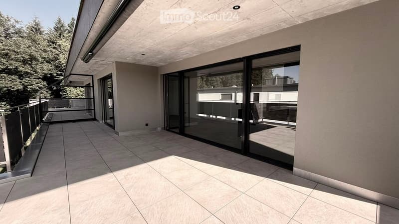 Modern balcony with large sliding glass doors, tiled floor, and metal railing, overlooking a landscaped area with trees.