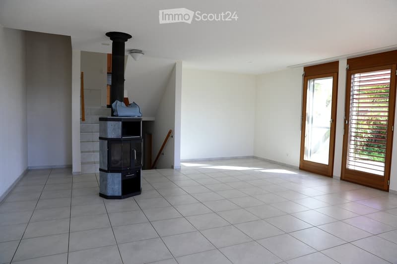 Empty room with tiled floor, white walls, and a modern wood-burning stove. There are two large sliding glass doors leading outside, and a staircase is visible in the background.