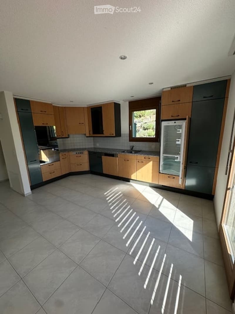 Modern kitchen with light wood cabinets, stainless steel appliances, and tiled flooring. Sunlight streams in through a window, casting shadows on the floor.