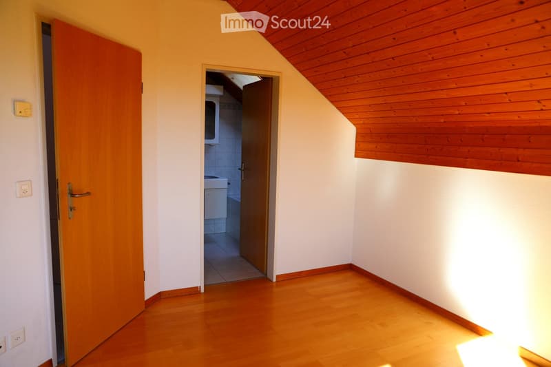 Empty hallway with wooden flooring, white walls, and a slanted wooden ceiling. An open door leads to a bathroom with a visible sink and bathtub.