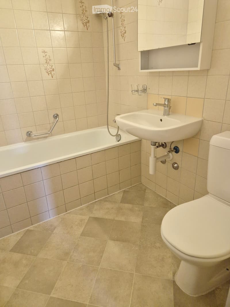 Bathroom with beige tiled walls and floor, featuring a bathtub, sink, toilet, and a small cabinet above the sink.