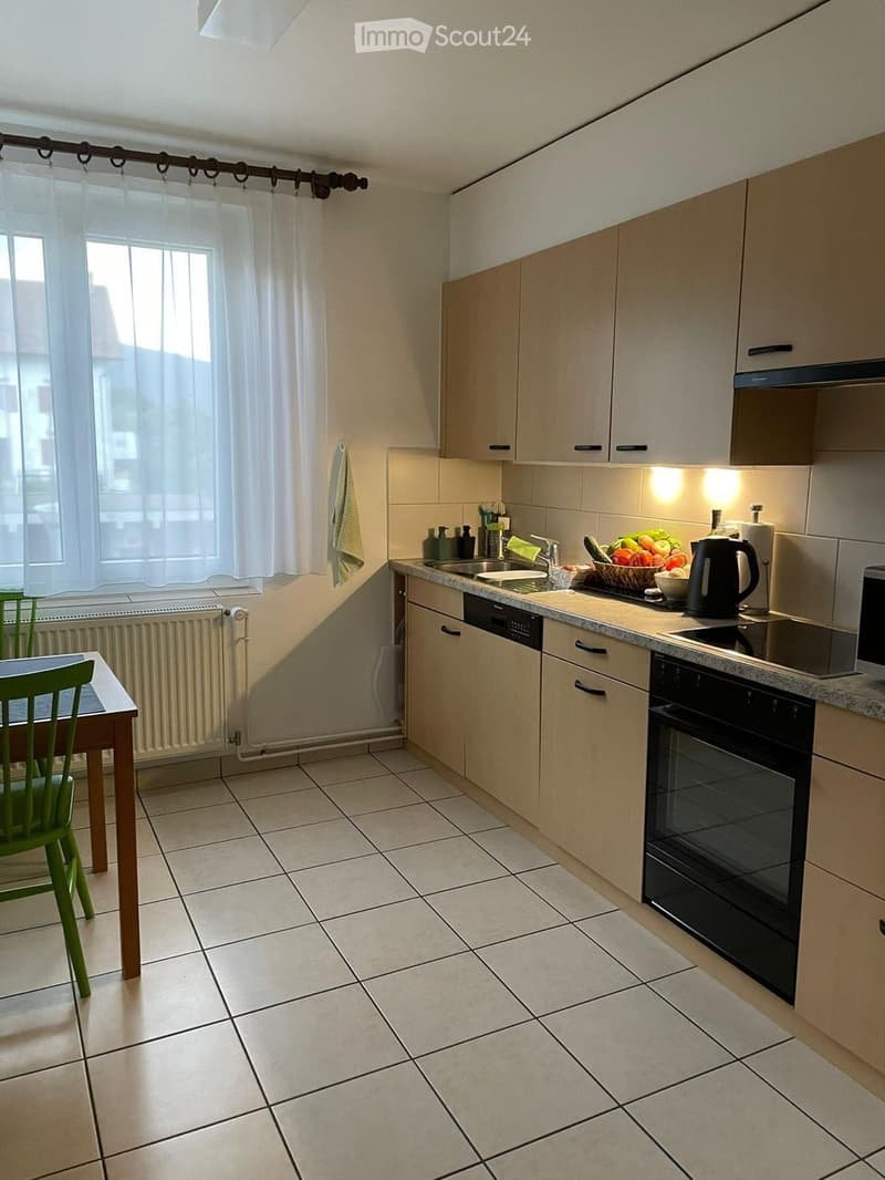 Modern kitchen with light wood cabinets, tiled floor, and various appliances including a sink, oven, microwave, and dishwasher. A small table with a green chair is placed near the window.
