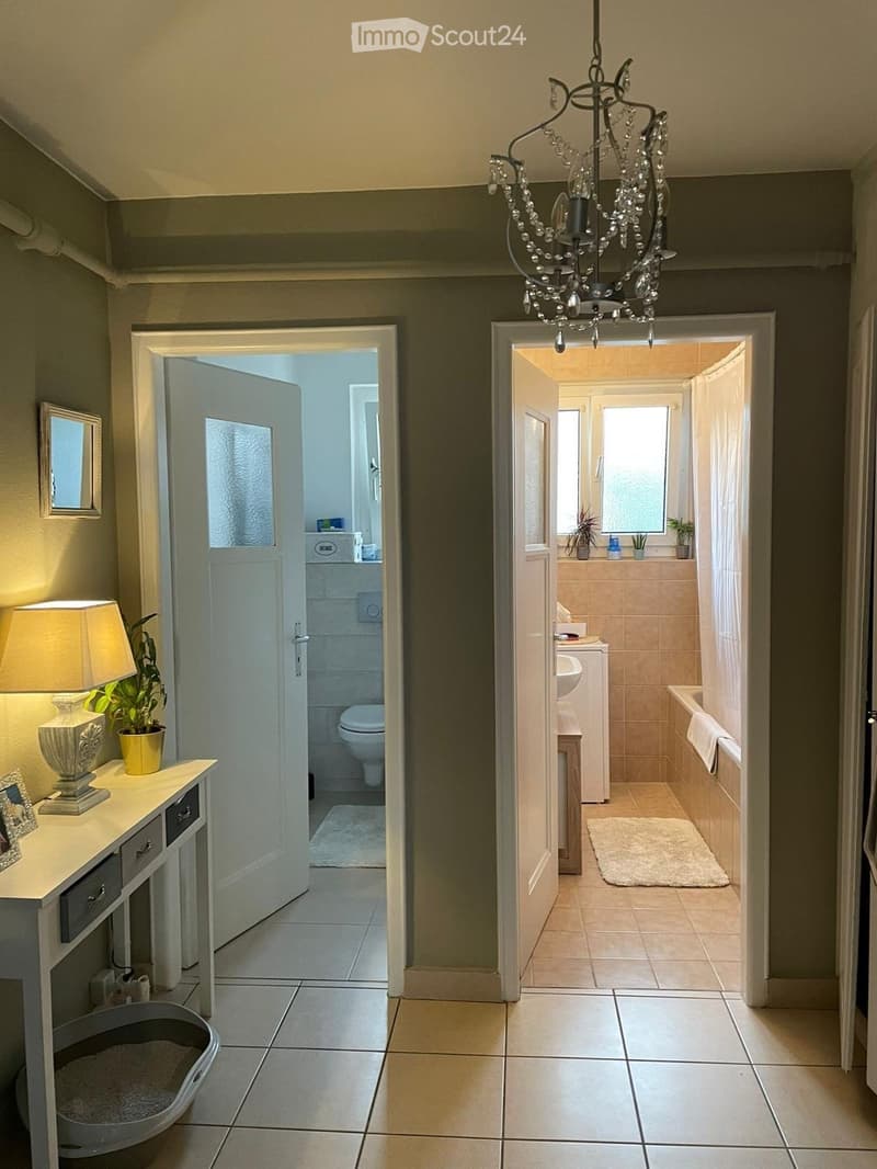 Hallway with tiled flooring, chandelier, two doors leading to a bathroom and another room, small table with lamp and plant, and a pet litter box.