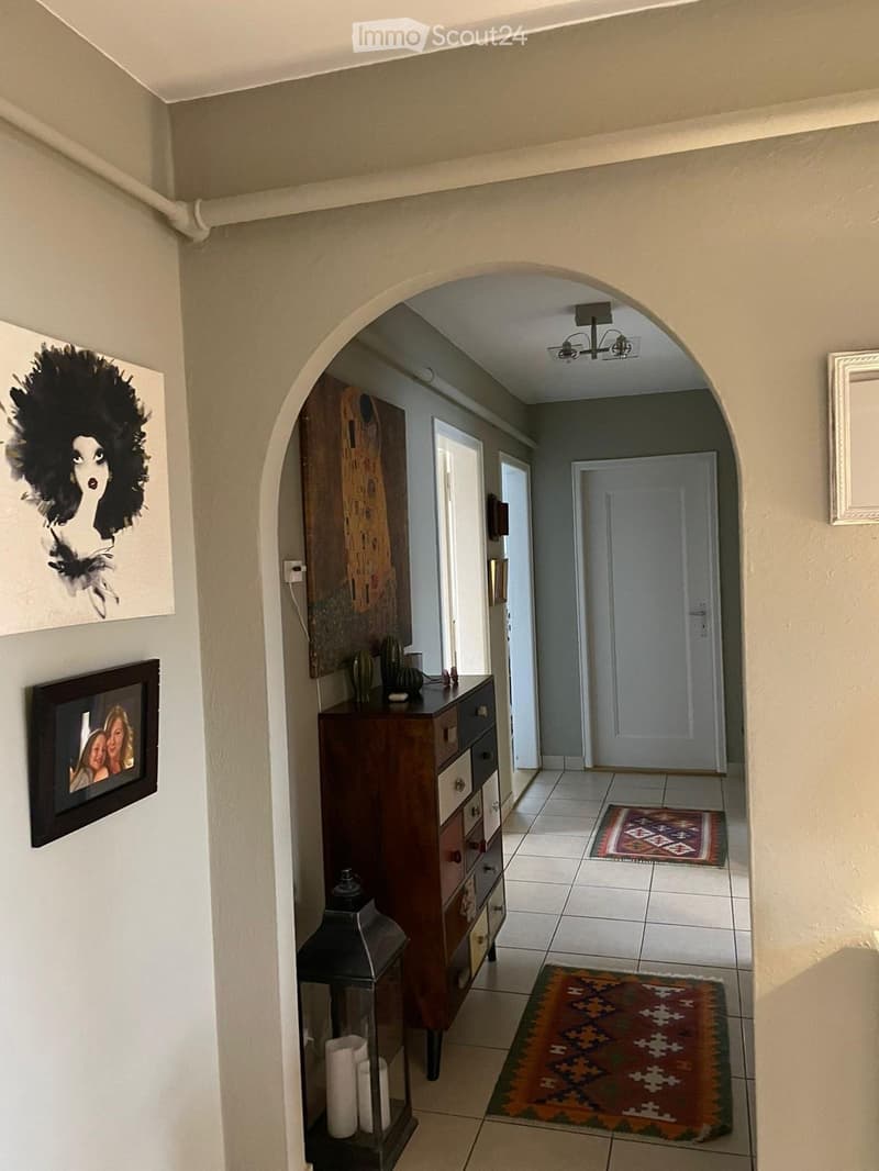 Hallway with beige walls, tiled floor, arched entryway, small wooden cabinet, decorative items, framed artwork, and patterned rugs.