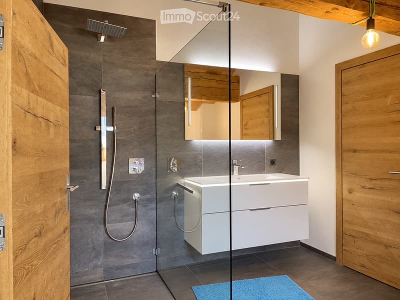 Modern bathroom with a glass-enclosed shower, grey tiles, white sink cabinet, and wooden accents.